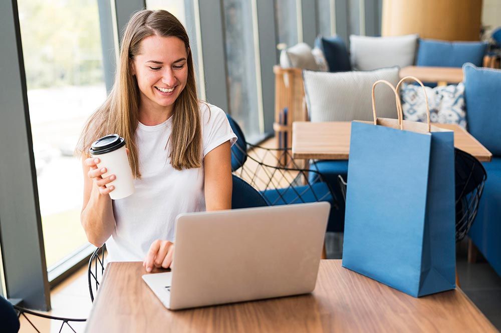 A woman is sitting at a table using a laptop computer while holding a cup of coffee.