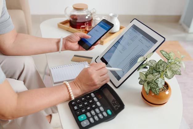 A woman is sitting at a table using a tablet and a cell phone.