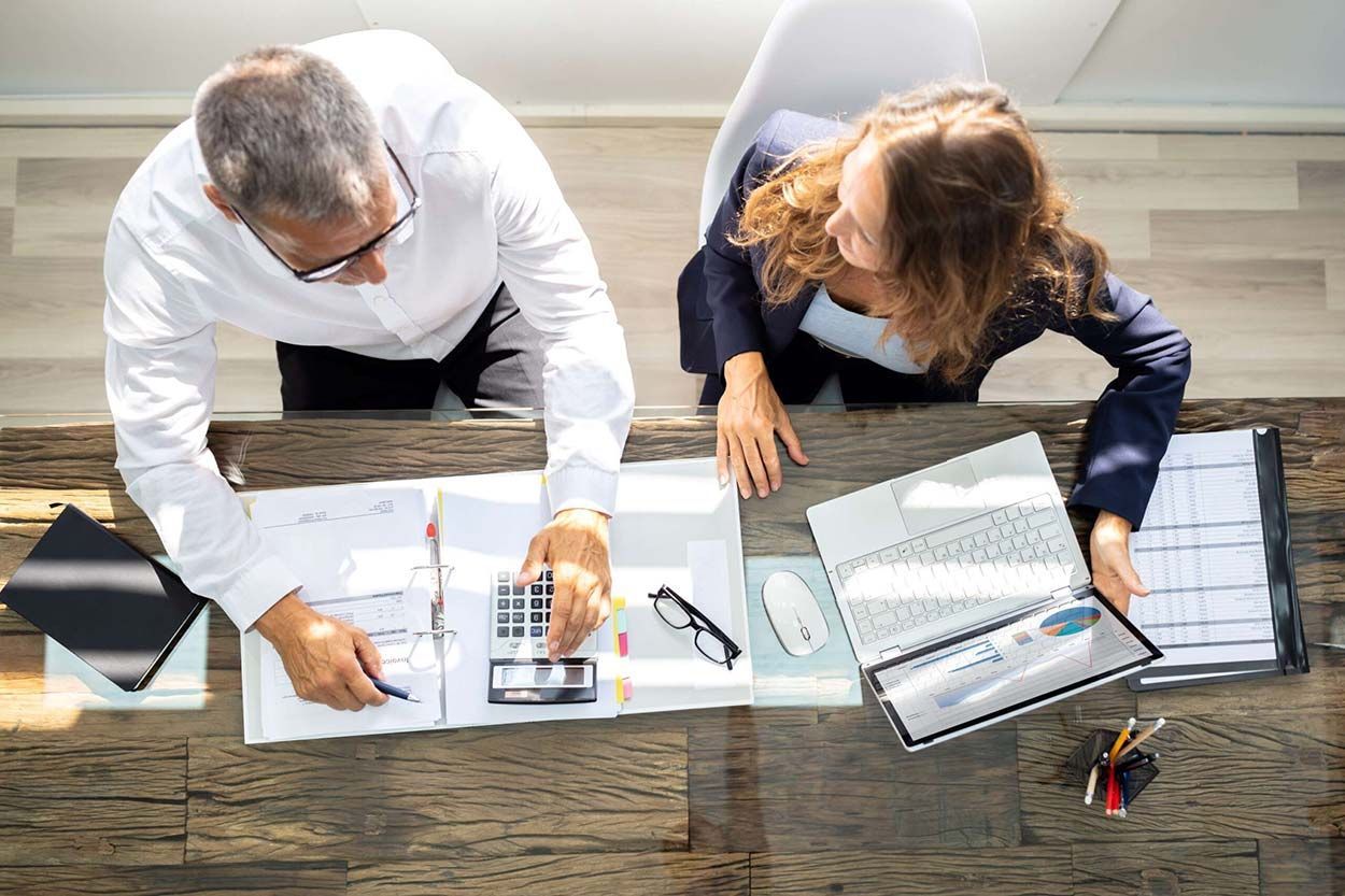 A man and a woman are sitting at a desk looking at papers and a calculator.