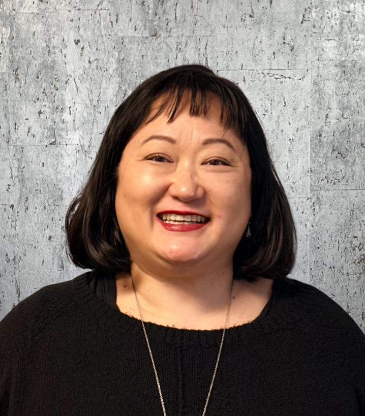 Woman with dark hair smiles, wearing a black top and a silver necklace, against a textured gray background.