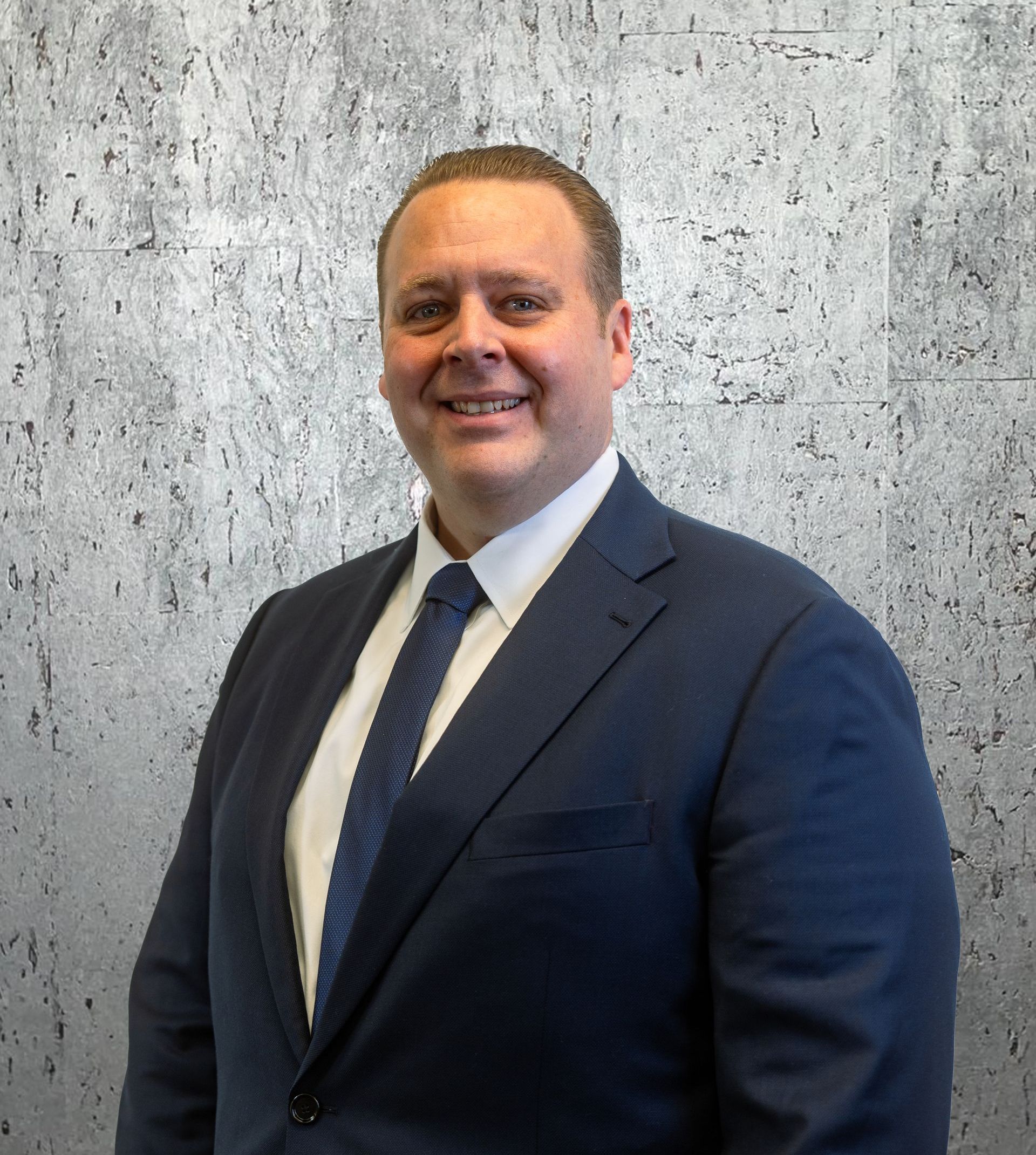 A man in a suit and tie is smiling in front of a concrete wall.