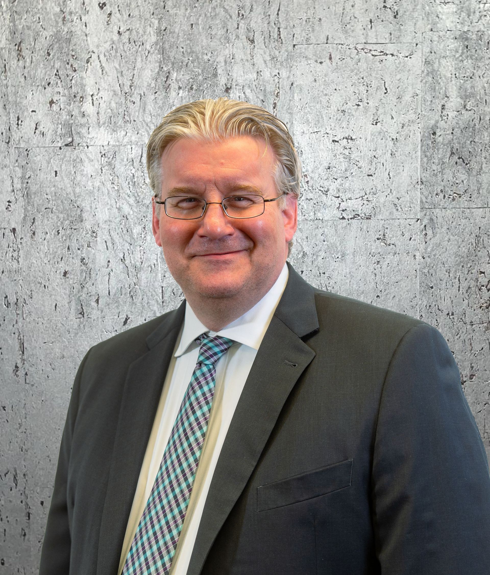A man in a suit and tie is smiling in front of a concrete wall.