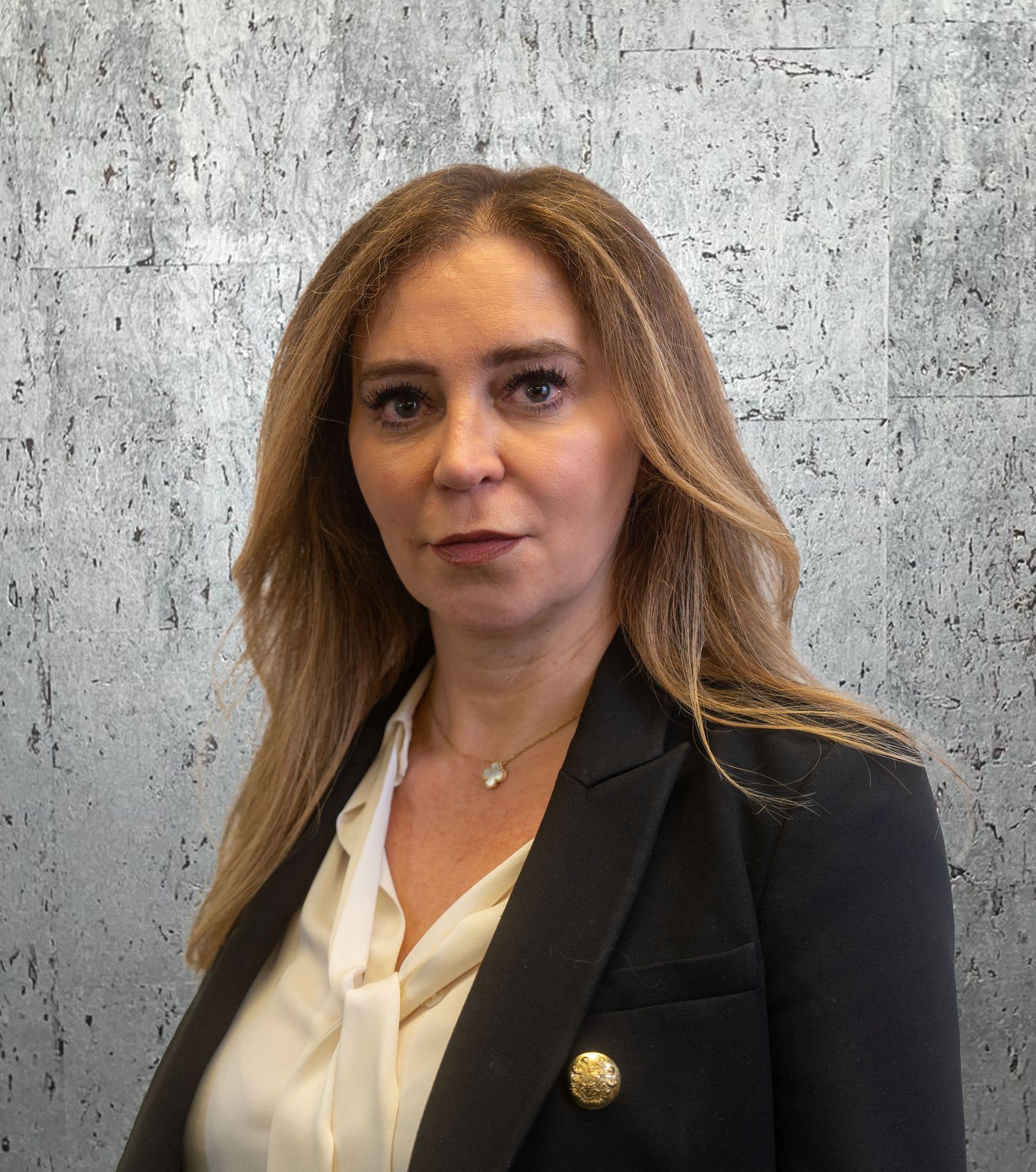 A woman wearing a black jacket and a white shirt is standing in front of a concrete wall.