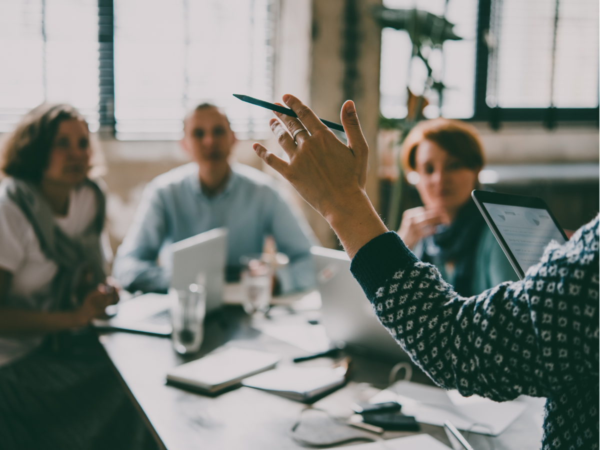 Person leading a meeting, gesturing with a pen, other people listening. Laptops and papers on the table.