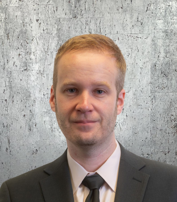 A man in a suit and tie stands in front of a concrete wall