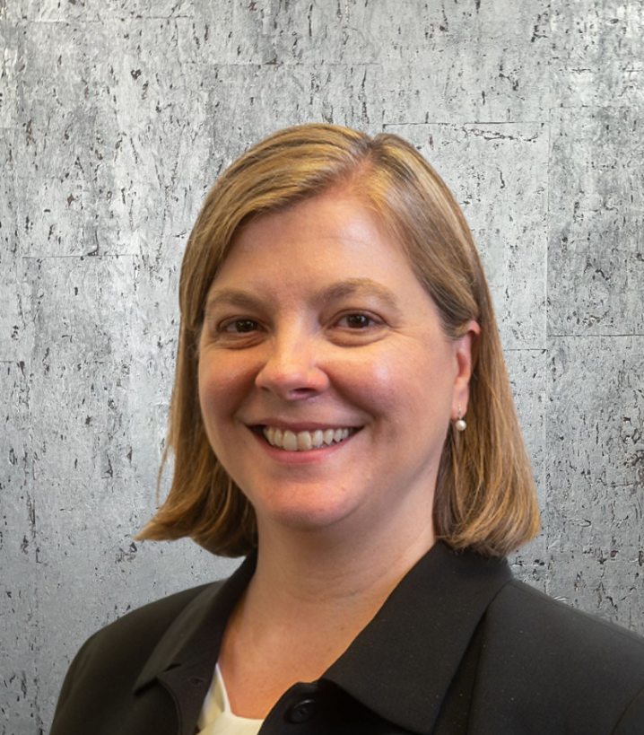 A woman in a black jacket is smiling in front of a concrete wall.