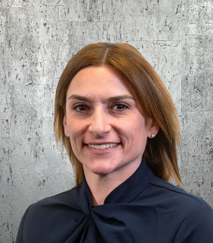 A woman in a blue shirt is smiling in front of a concrete wall.