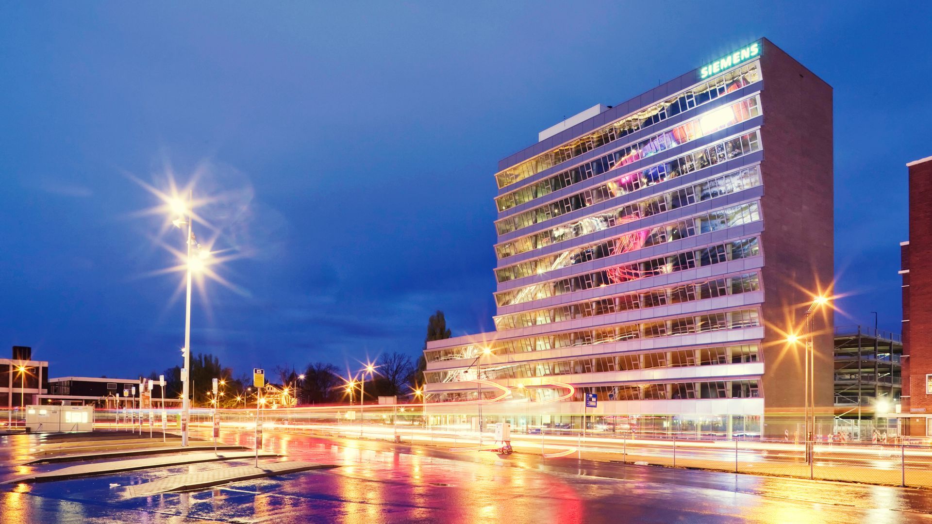 Modern kantoorgebouw 's nachts met spiegelende ramen, straatverlichting en een plein dat glimt van de regen en de lampen weerkaatst