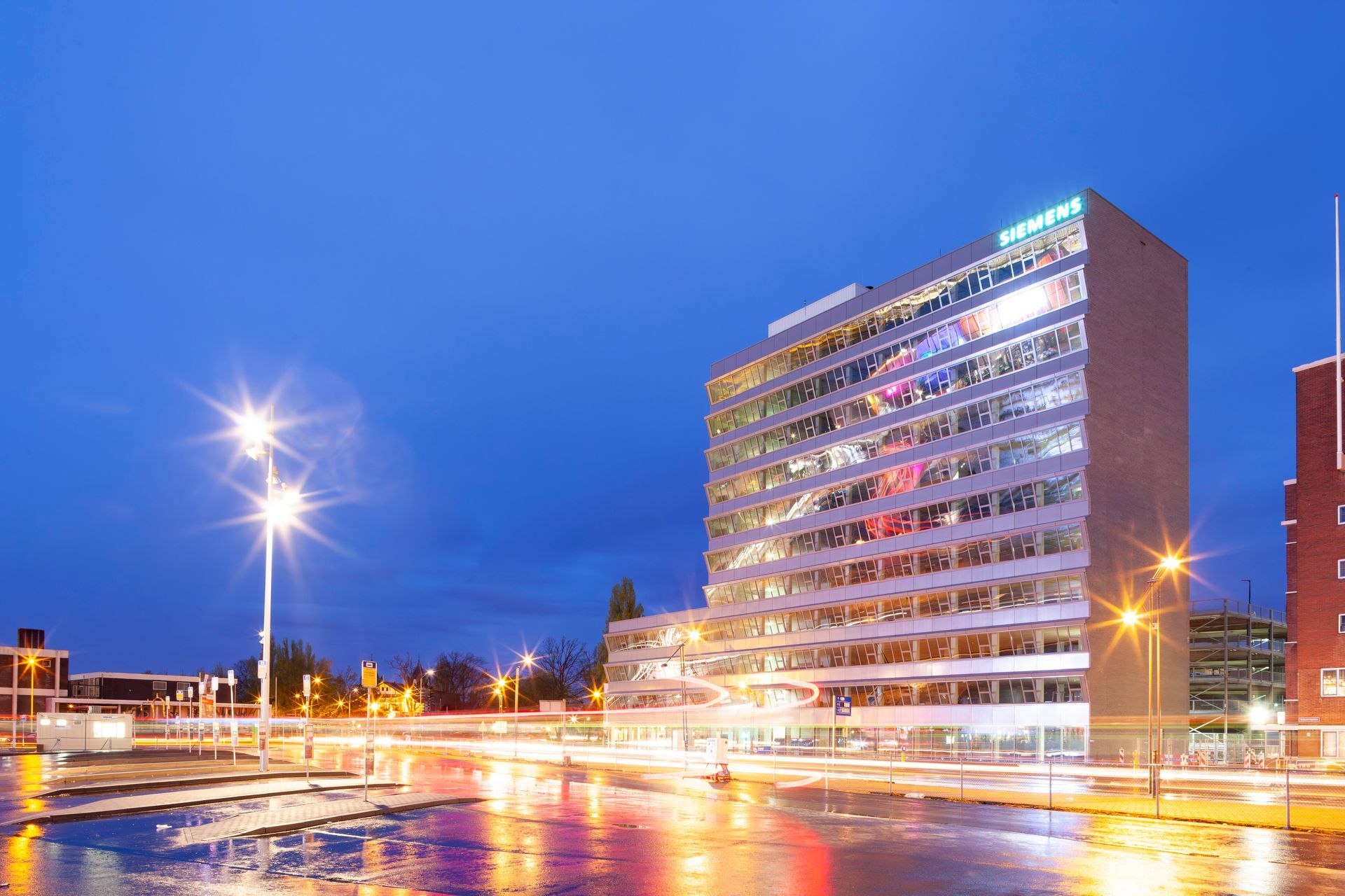 Modern kantoorgebouw in de schemering met verlichte ramen, weerspiegeld in een natte straat.