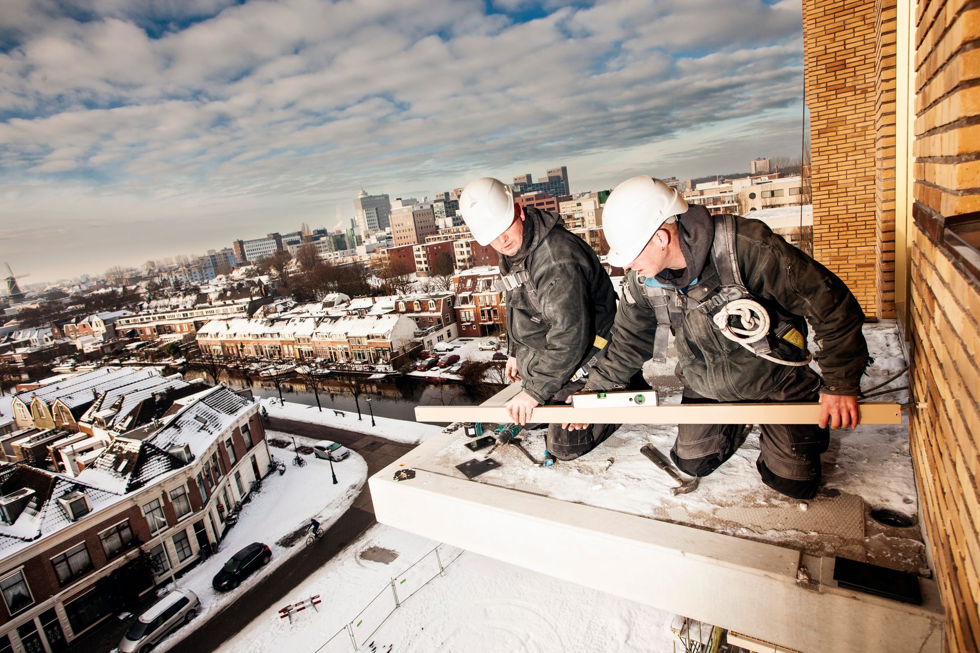 Twee bouwvakkers in veiligheidsuitrusting meten op grote hoogte op een balkon zonder leuning, met uitzicht op een besneeuwde stad. 