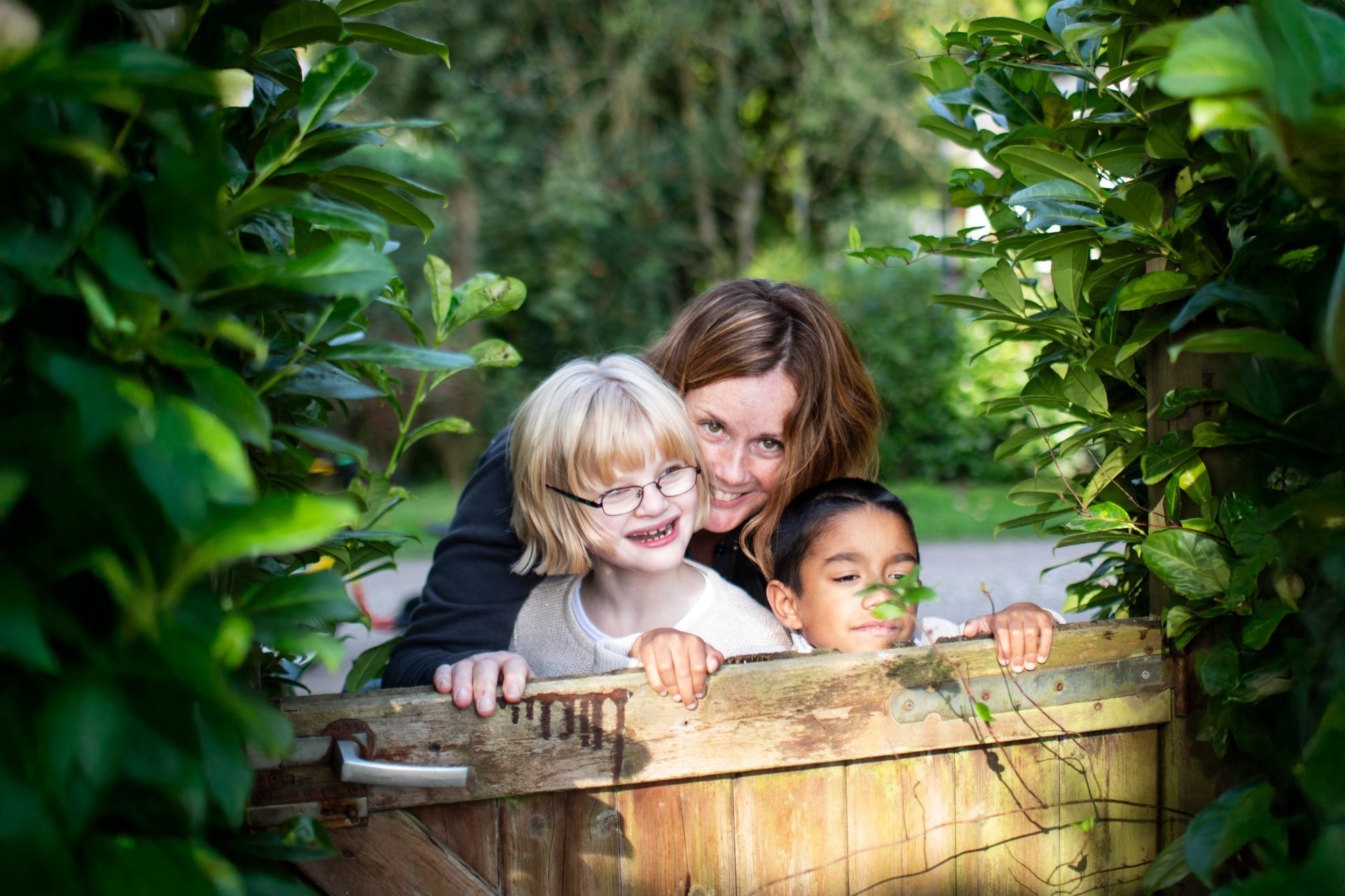 Een vrouw en twee kinderen glimlachen vanachter een houten hek, omringd door groen gebladerte.