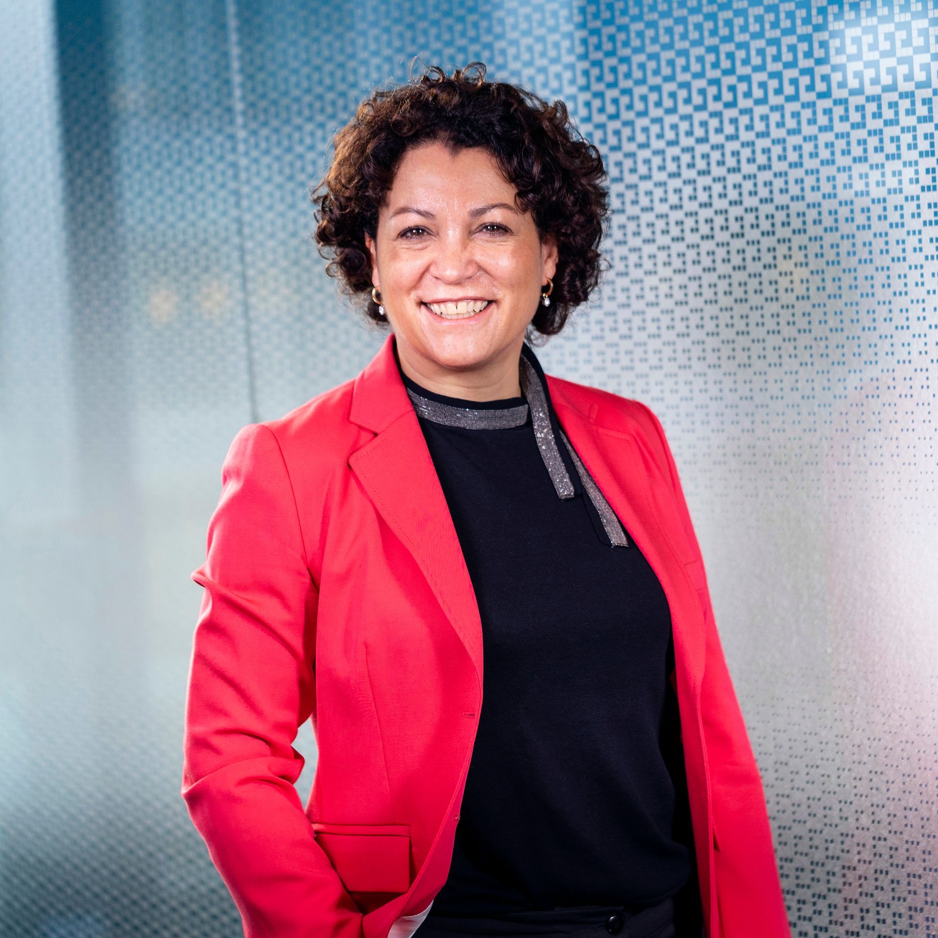 Een vrouw in een rode blazer glimlacht, staand tegen een glazen wand met een patroon.