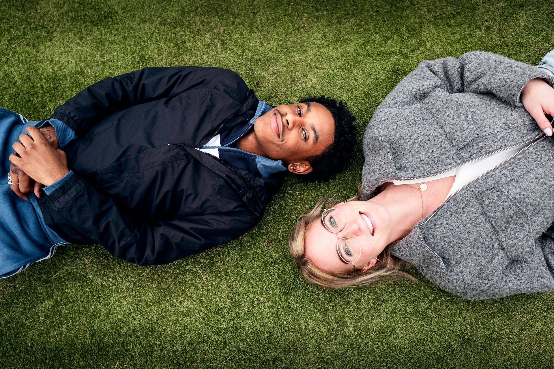 Twee mensen liggen op hun rug in het gras en kijken omhoog. De een draagt ​​een blauwe jas, de ander een grijze mantel.