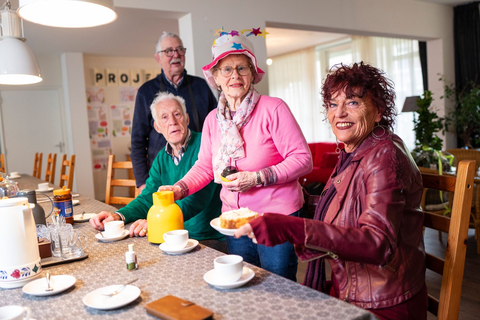 Een groepje van vier senioren zit aan een tafel, twee van hen houden taart vast. Een van hen draagt ​​een feestmuts, ze glimlachen allemaal in een lichte ruimte.