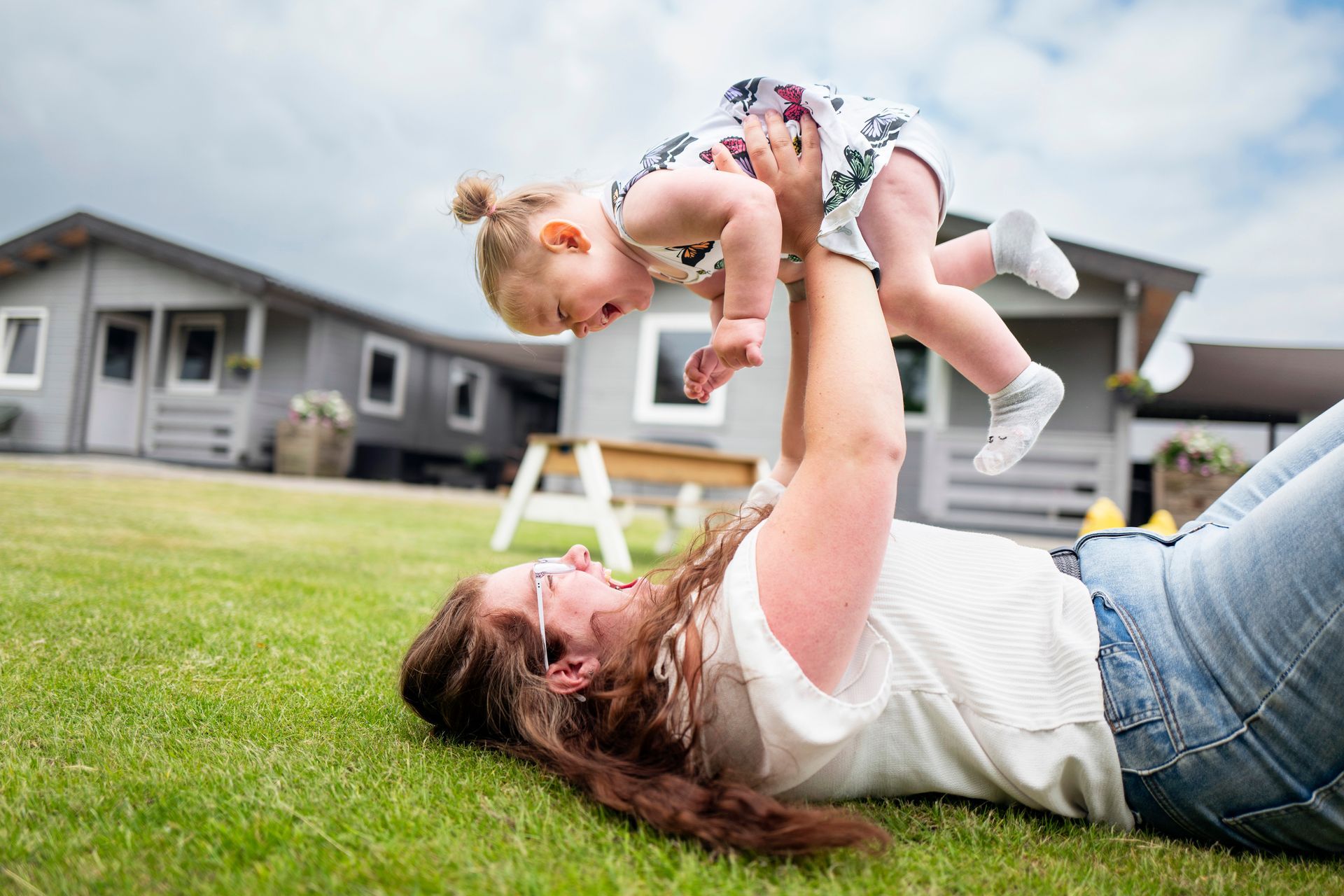 Jonge moeder ligt op haar rug in het gras, tilt een kind op en lacht. Huisjes op de achtergrond.