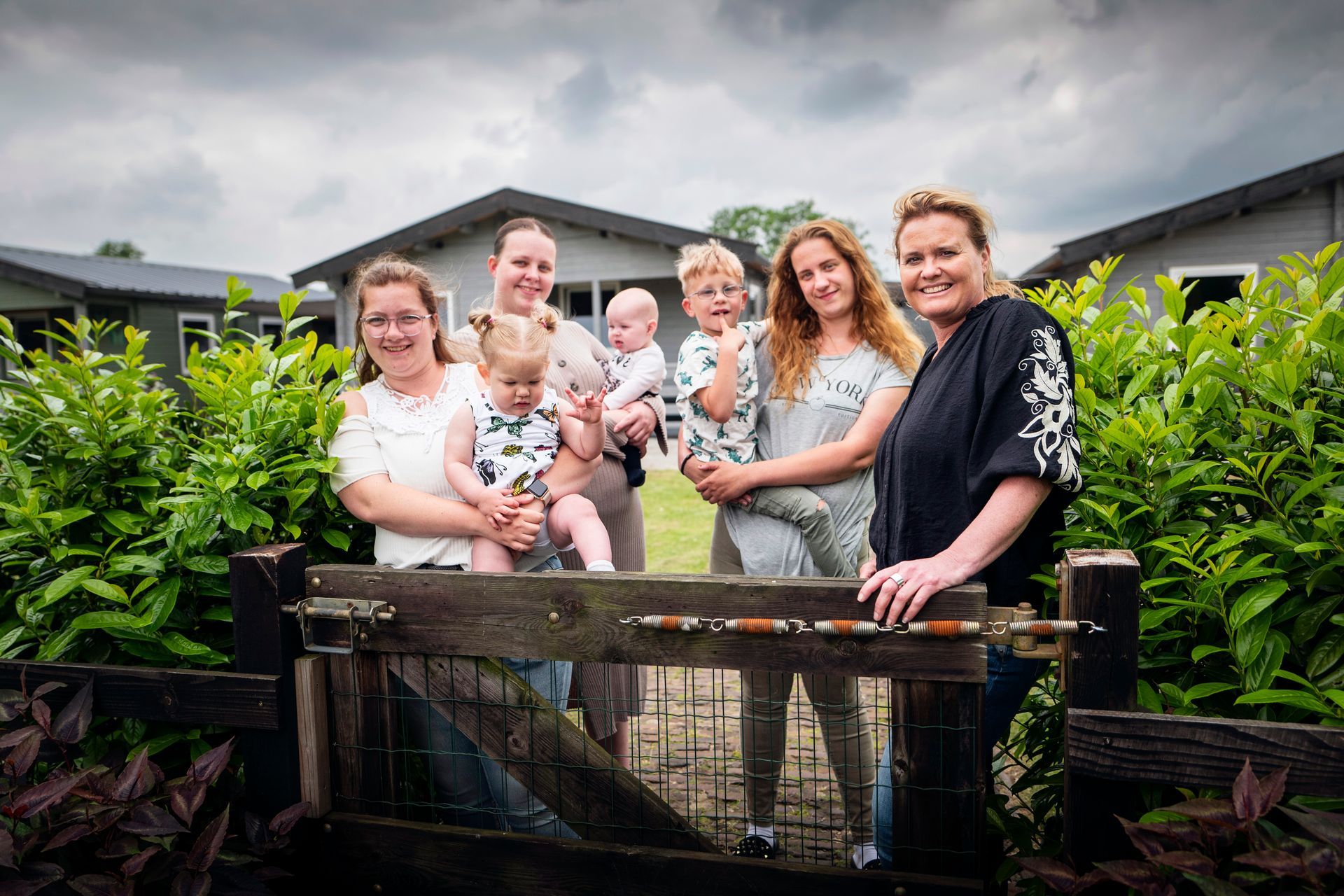 Een groep van vier jonge vrouwen met jonge kinderen voor een houten hek, struiken en een huisjes op de achtergrond.