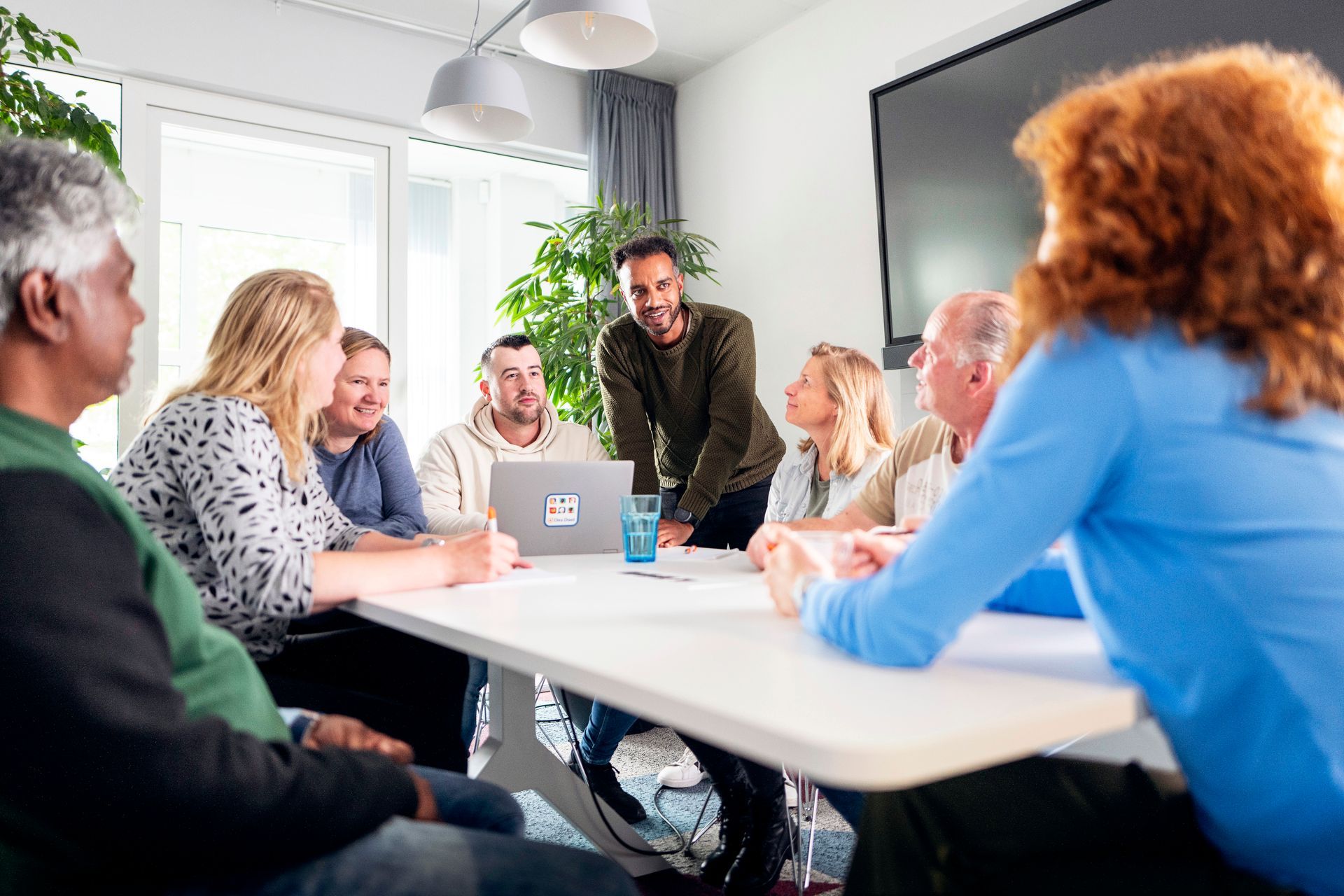 Mensen zitten in een vergadering rond een tafel, één van hen staat te spreken.