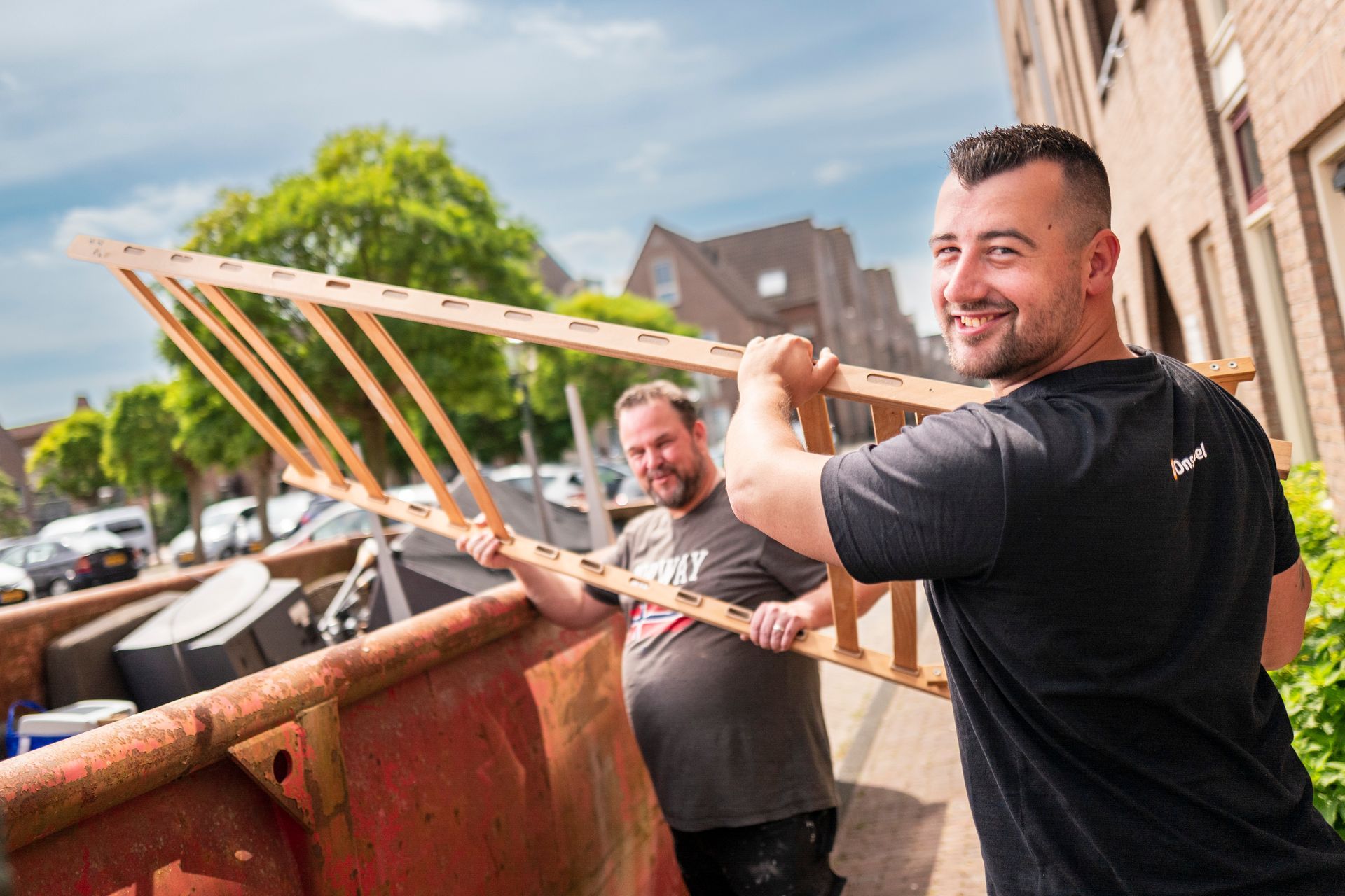 Twee mannen tillen een lattenbodem in een afvalcontainer in een stedelijke omgeving. Een van de mannen glimlacht terwijl hij de lattenbodem vasthoudt.