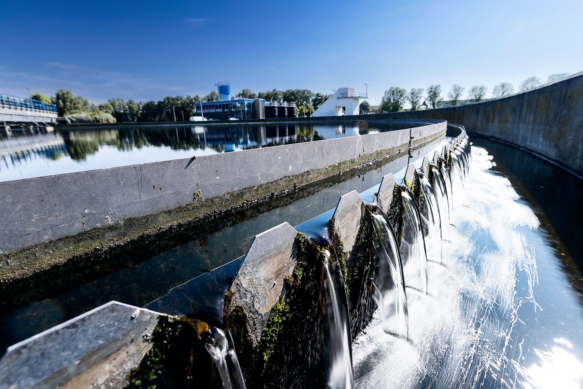 Waterzuiveringsinstallatie met betonnen constructie en stromend water. Blauwe lucht op de achtergrond en gespiegeld in het water.