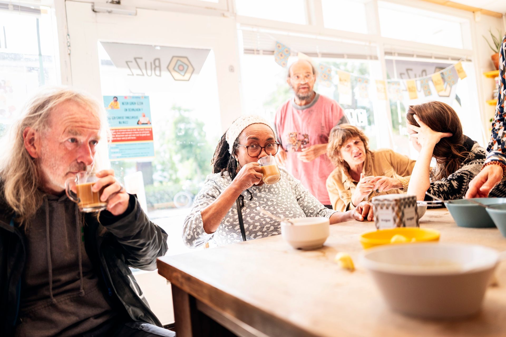 Mensen zitten binnen in een buurthuis aan een tafel en drinken uit mokken. Achter hen staat een man in een roze shirt.