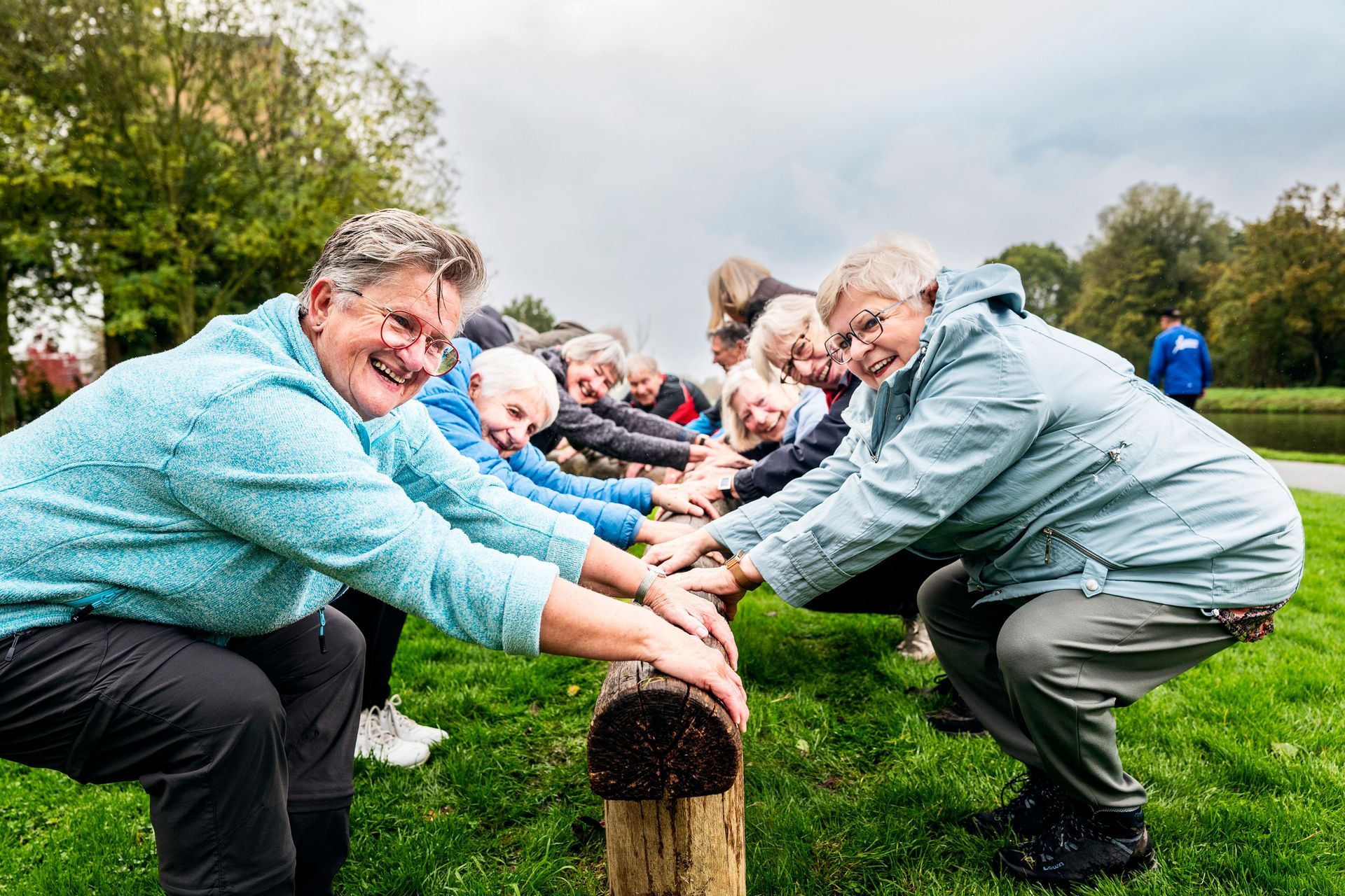 Een groep sportende oudere mensen in lichtblauwe jassen, lachend, houdt zich vast aan een houten balk in een park.