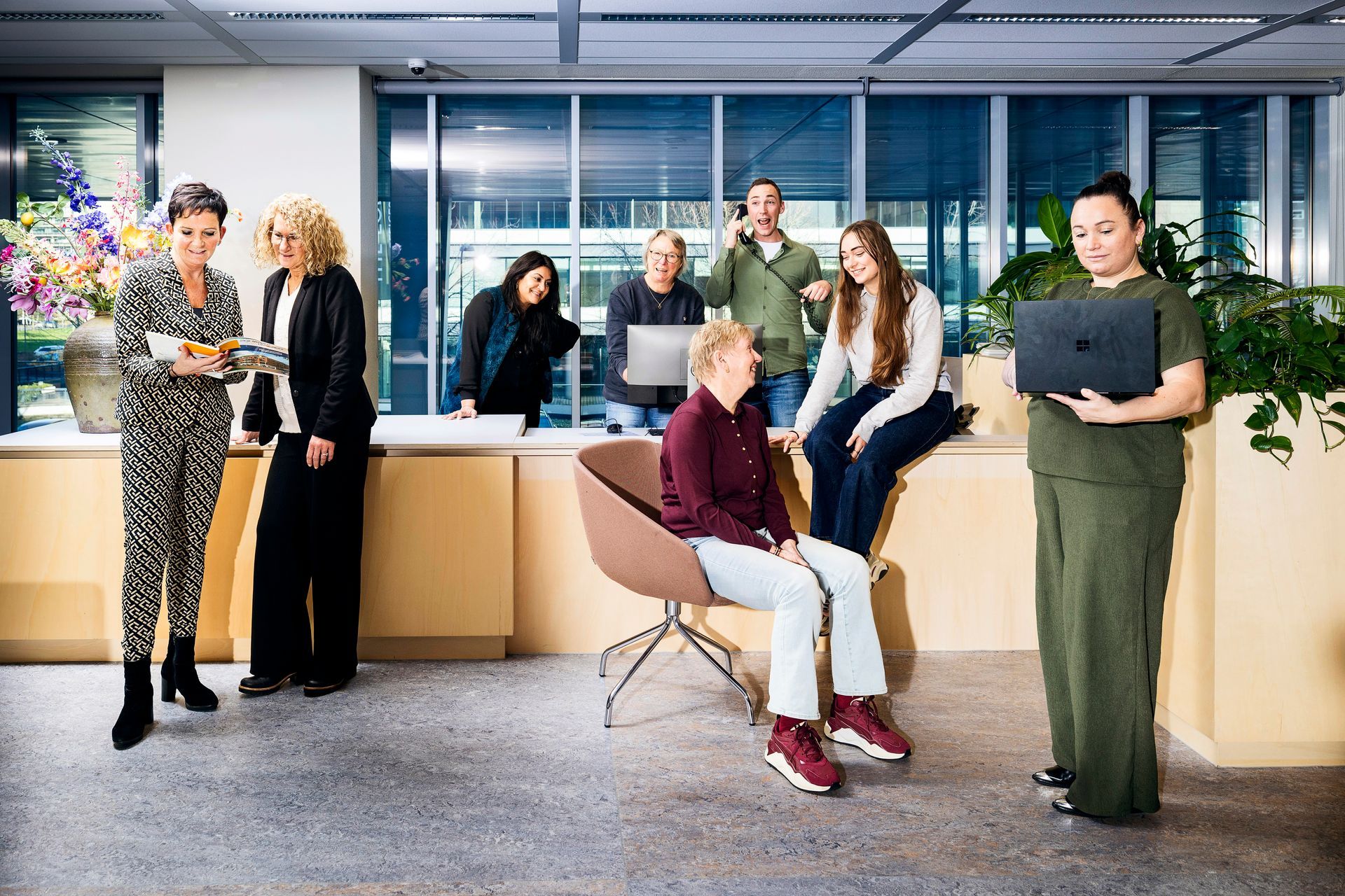 Een groep mensen in een kantooromgeving, sommigen aan het werk, anderen aan het praten, één persoon houdt een laptop vast.