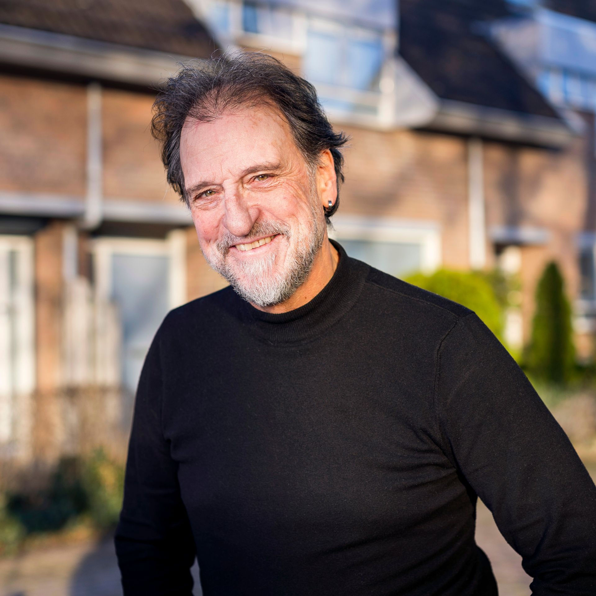 Een man met een grijzende baard, gekleed in een zwarte coltrui, glimlacht, buiten.