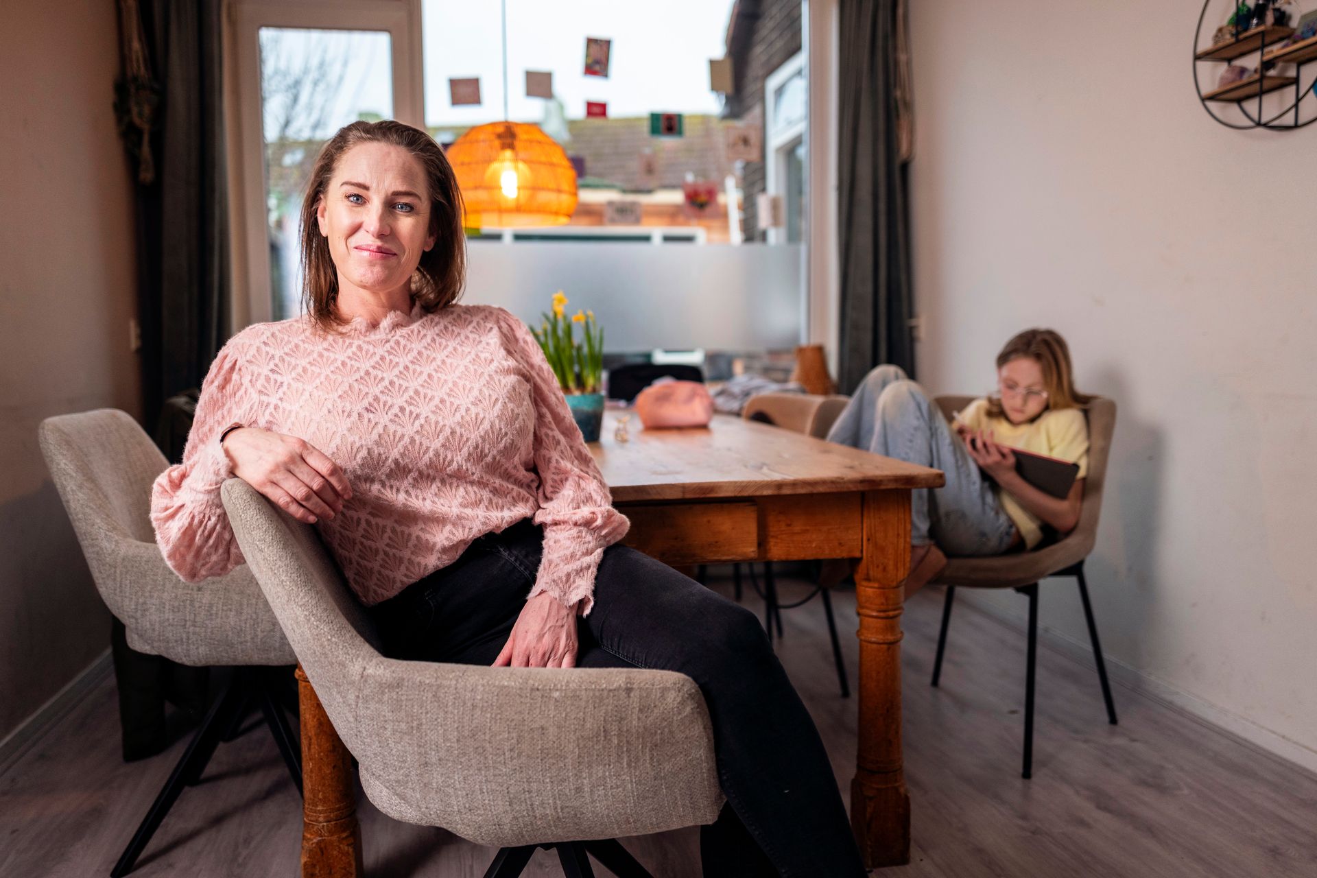 Een vrouw in een roze trui zit glimlachend aan een tafel. Op de achtergrond is een kind te zien dat een tablet gebruikt. Interieur.