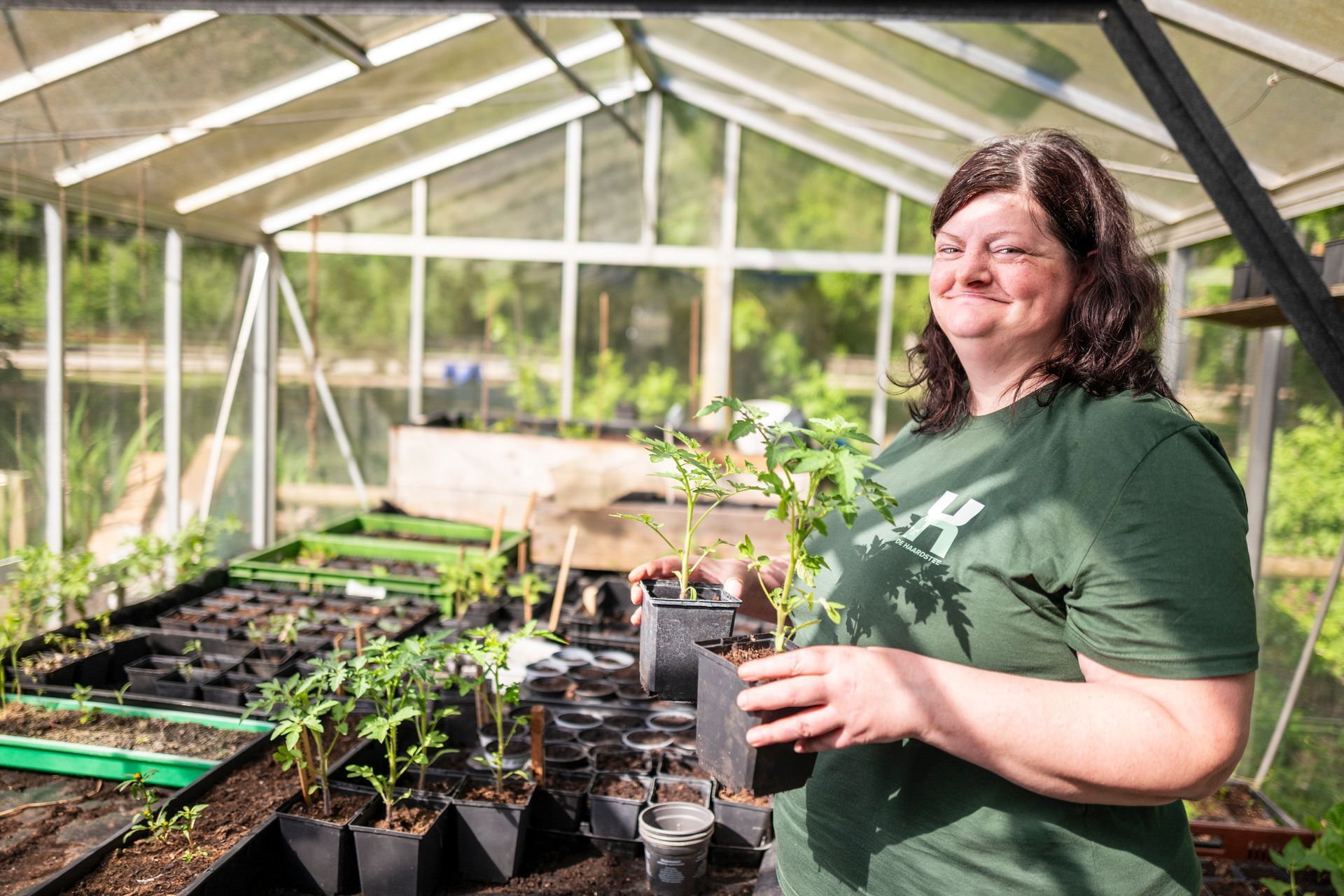 Een vrouw in een groene blouse lacht in een kas, terwijl ze een potplant tussen de zaailingen vasthoudt.