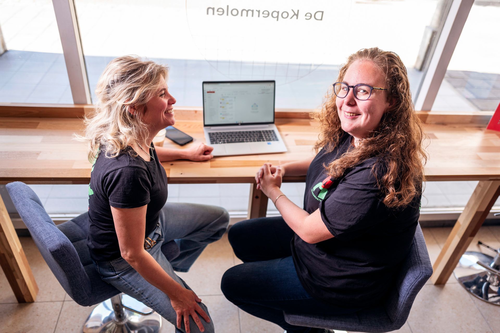Twee vrouwen glimlachen naar elkaar, zittend aan een bureau met een laptop, bij een raam.