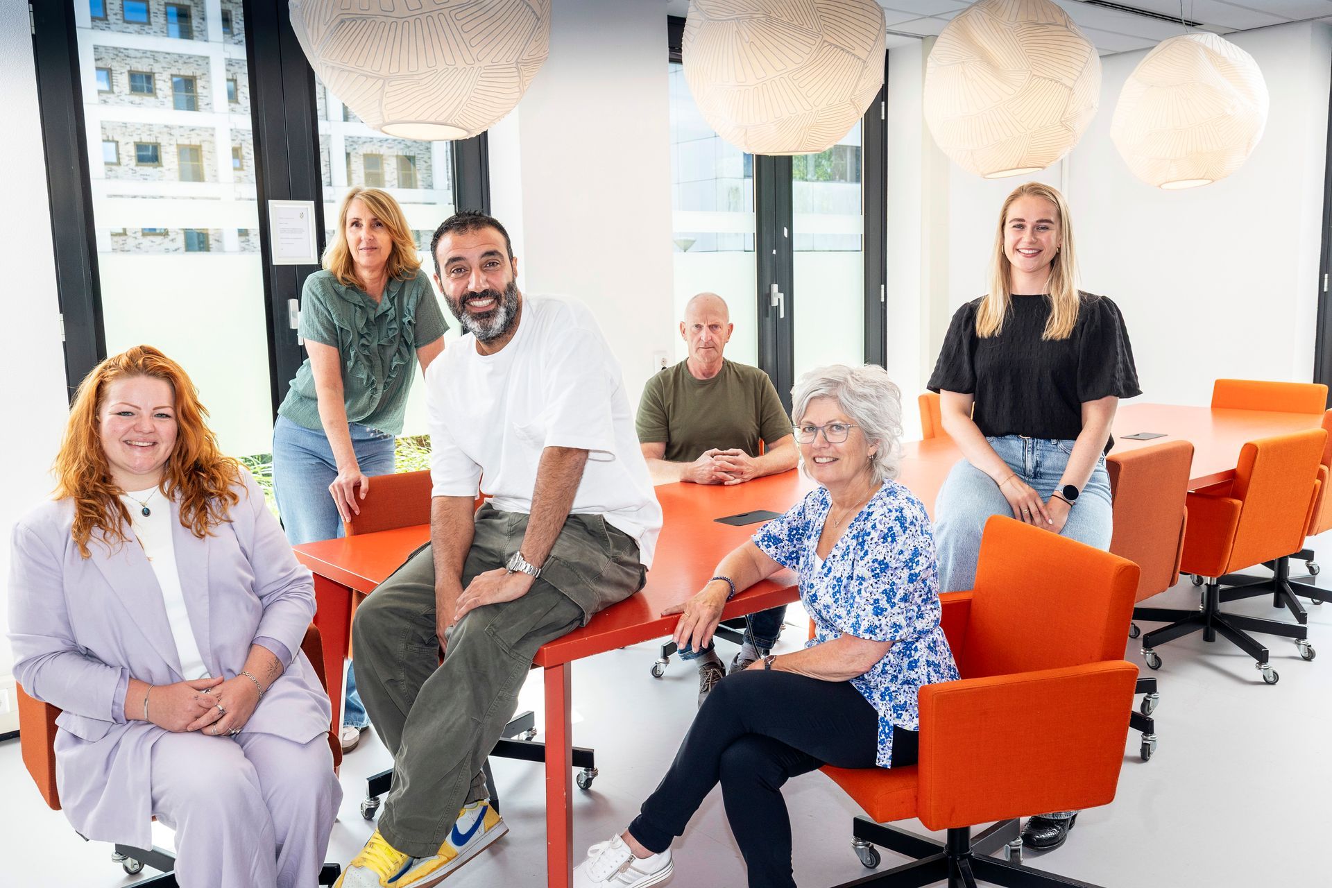 Een groep mensen zit in een licht kantoor rond een oranje tafel en lacht.