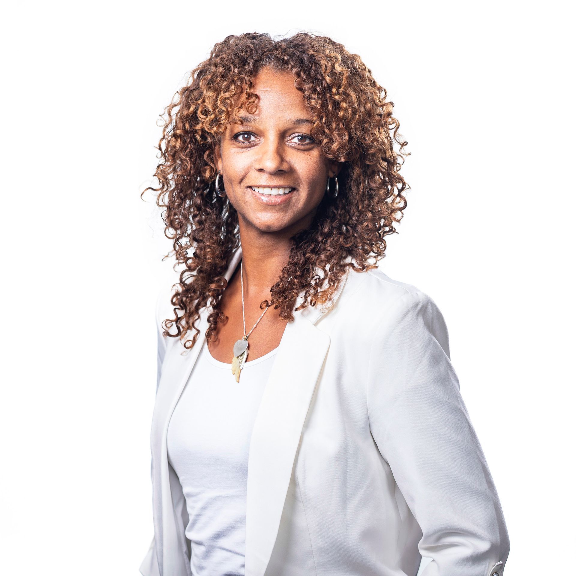 Vrouw met krullend bruin haar, gekleed in een witte blazer en met een glimlach.