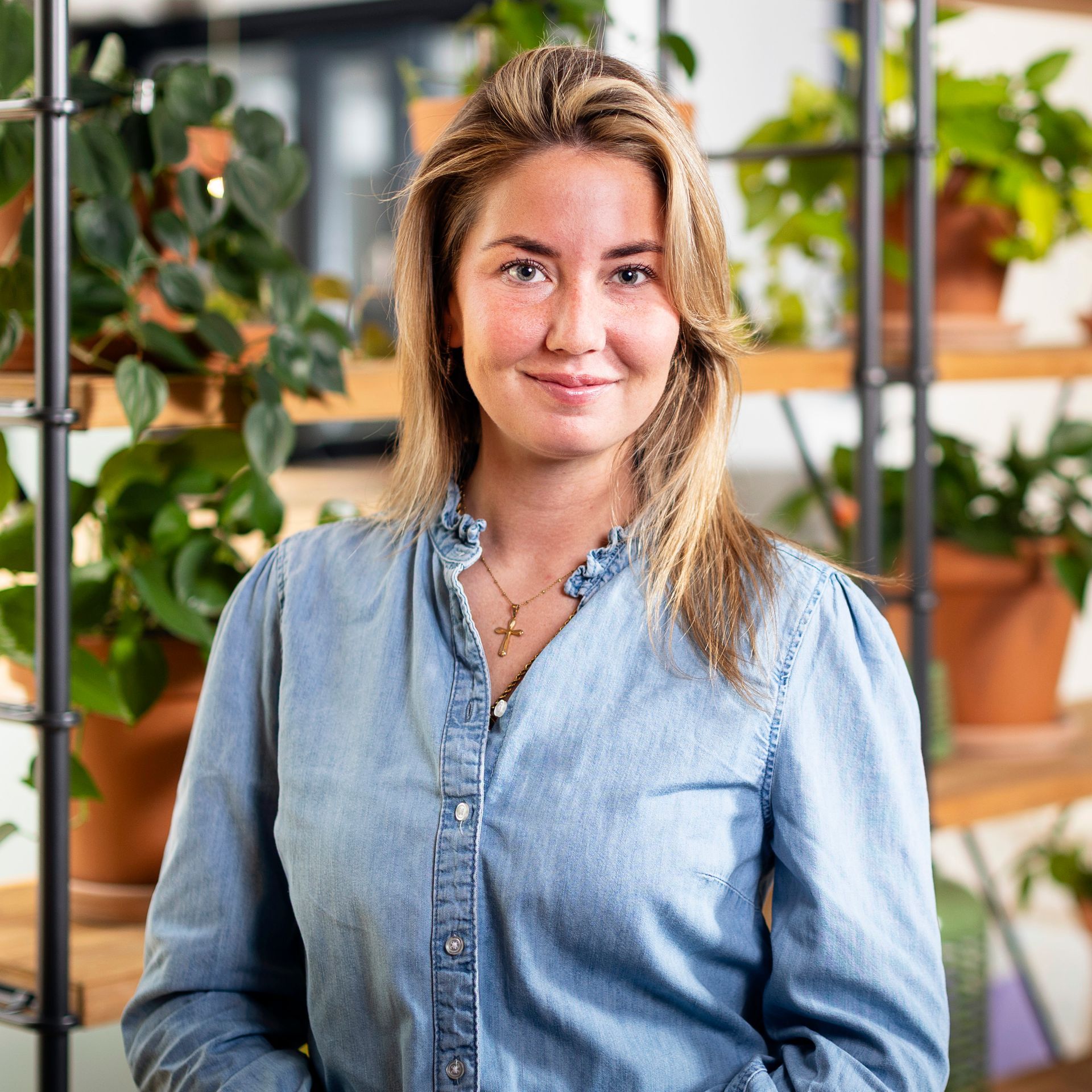 Een vrouw in een spijkerblouse lacht terwijl ze voor een schap met potplanten staat.