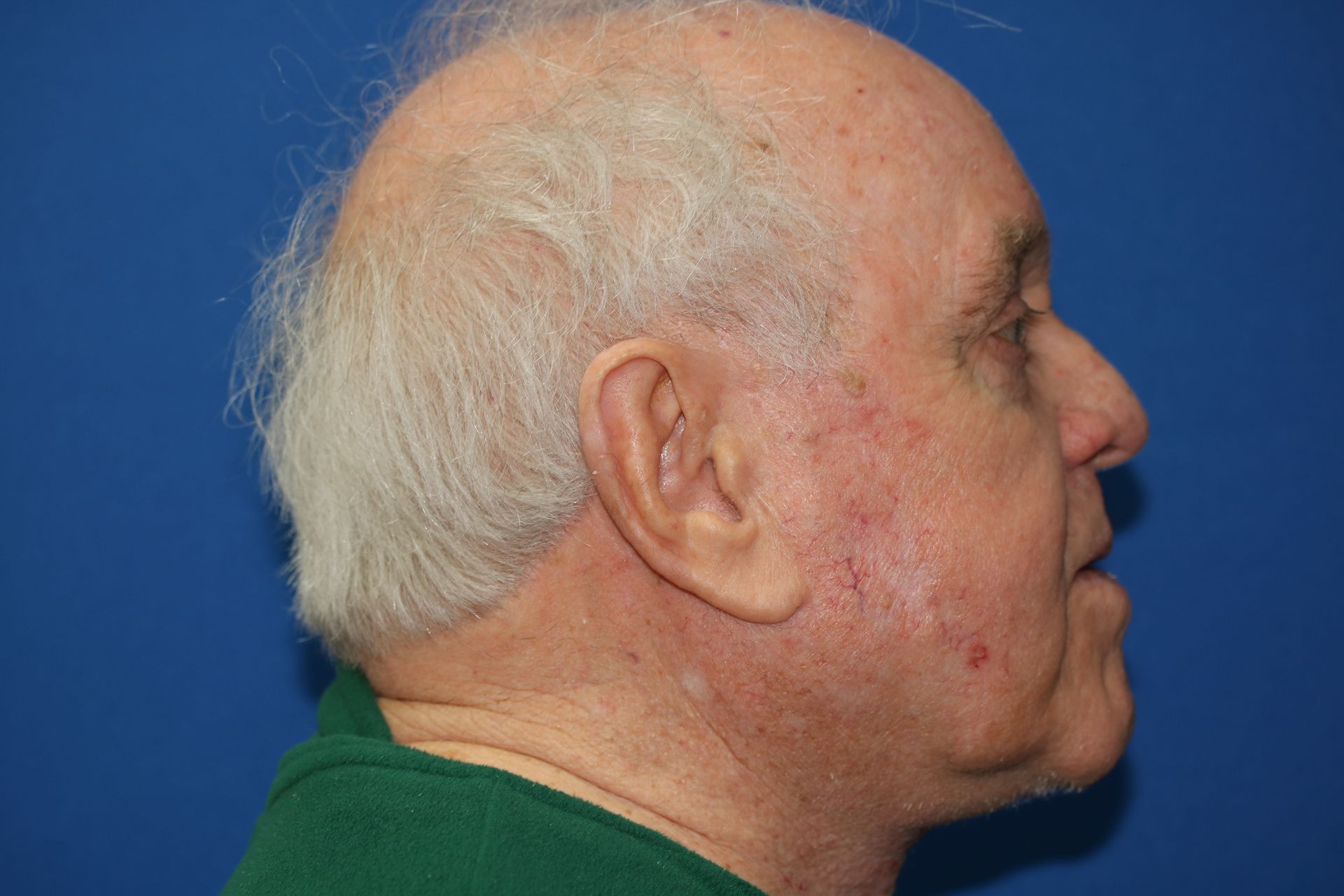 A man with gray hair and a green shirt is standing in front of a blue wall.