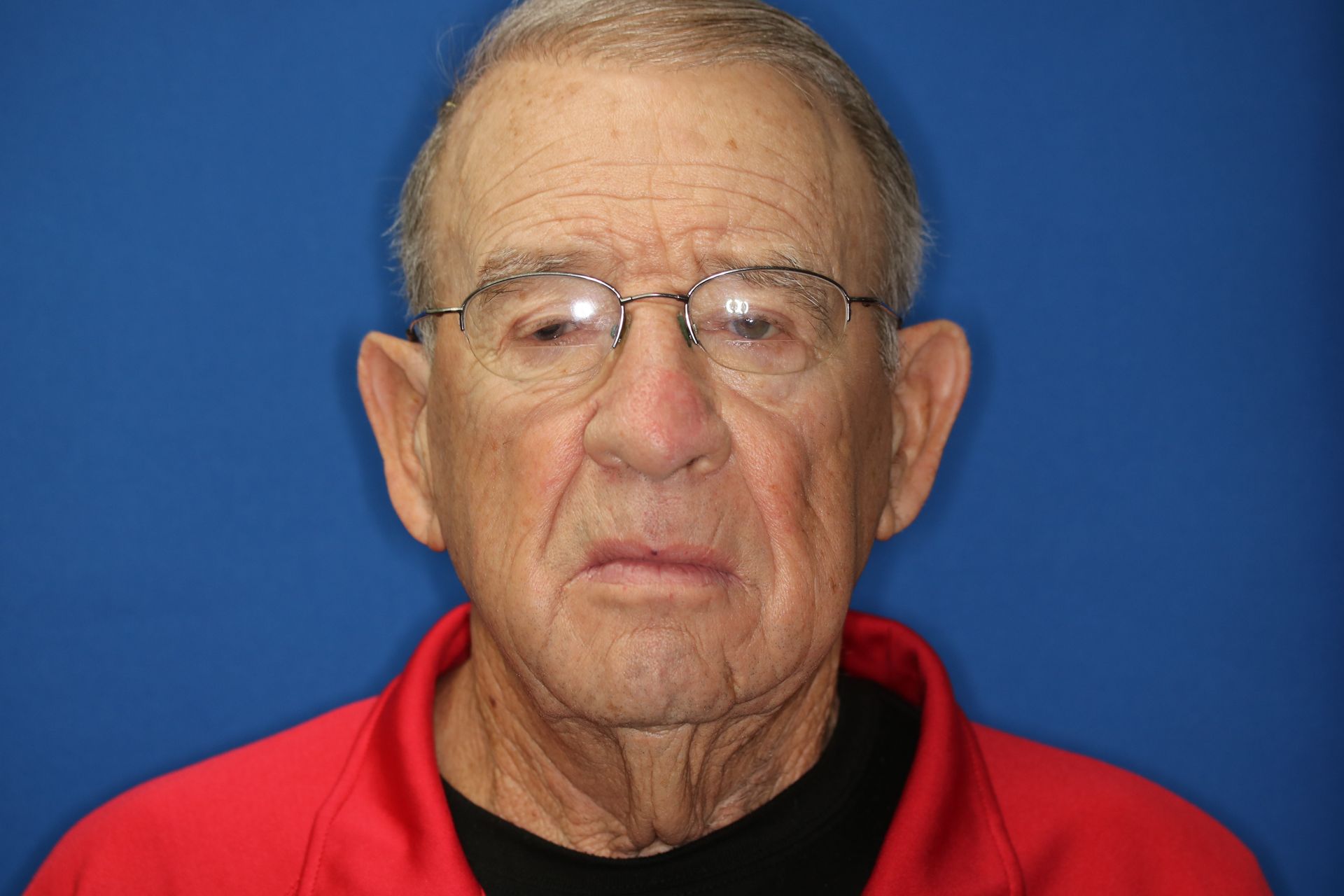 An older man wearing glasses and a red shirt is standing in front of a blue background.