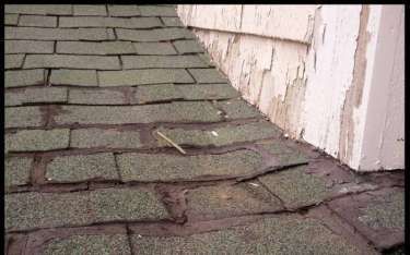 Damaged asphalt shingles on a roof corner next to peeling white siding.