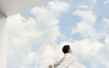 Person looking up, reaching towards a bright blue sky with fluffy white clouds, framed by a white wall.