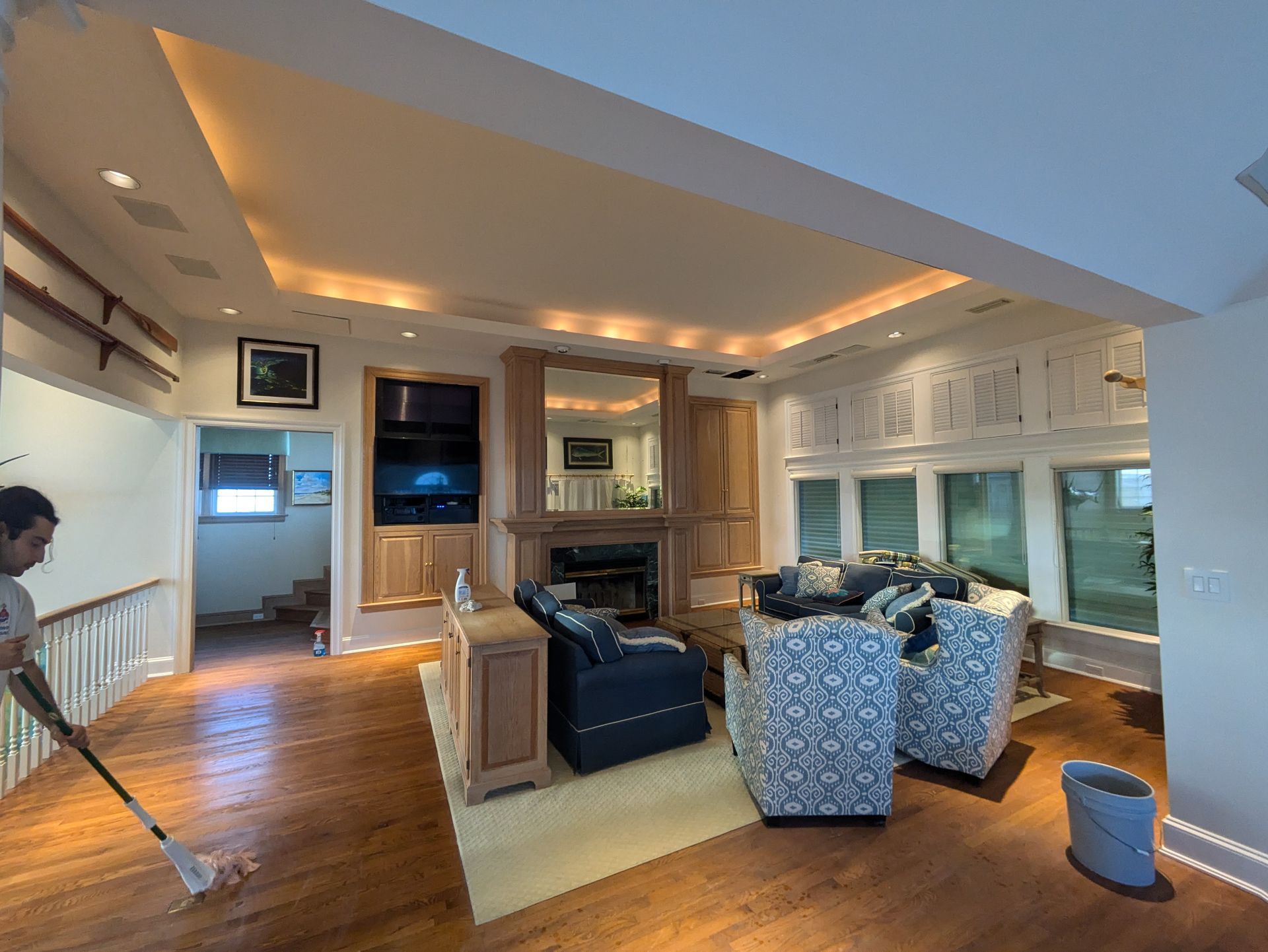 Man mopping wooden floor in a living room with couches, fireplace, and built-ins.