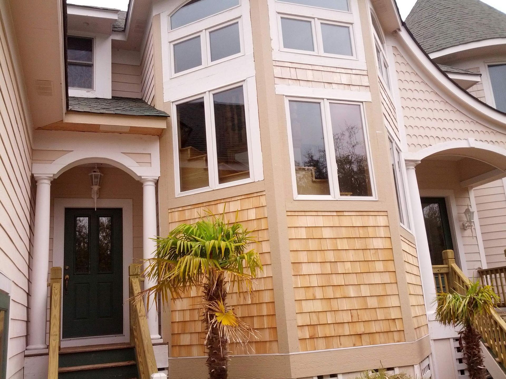 Two-story house with light brown wood shingles, beige siding, large windows, and a green front door.