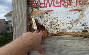 Hand touching rotted wood siding with damaged building wrap, revealing underlying rot, outdoors.