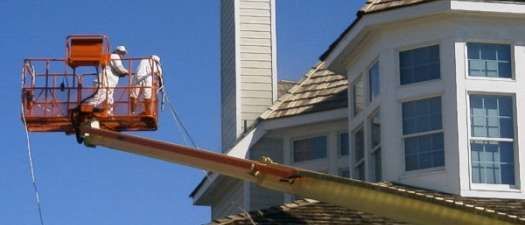 Workers in lift painting the exterior of a white house with a blue sky background.