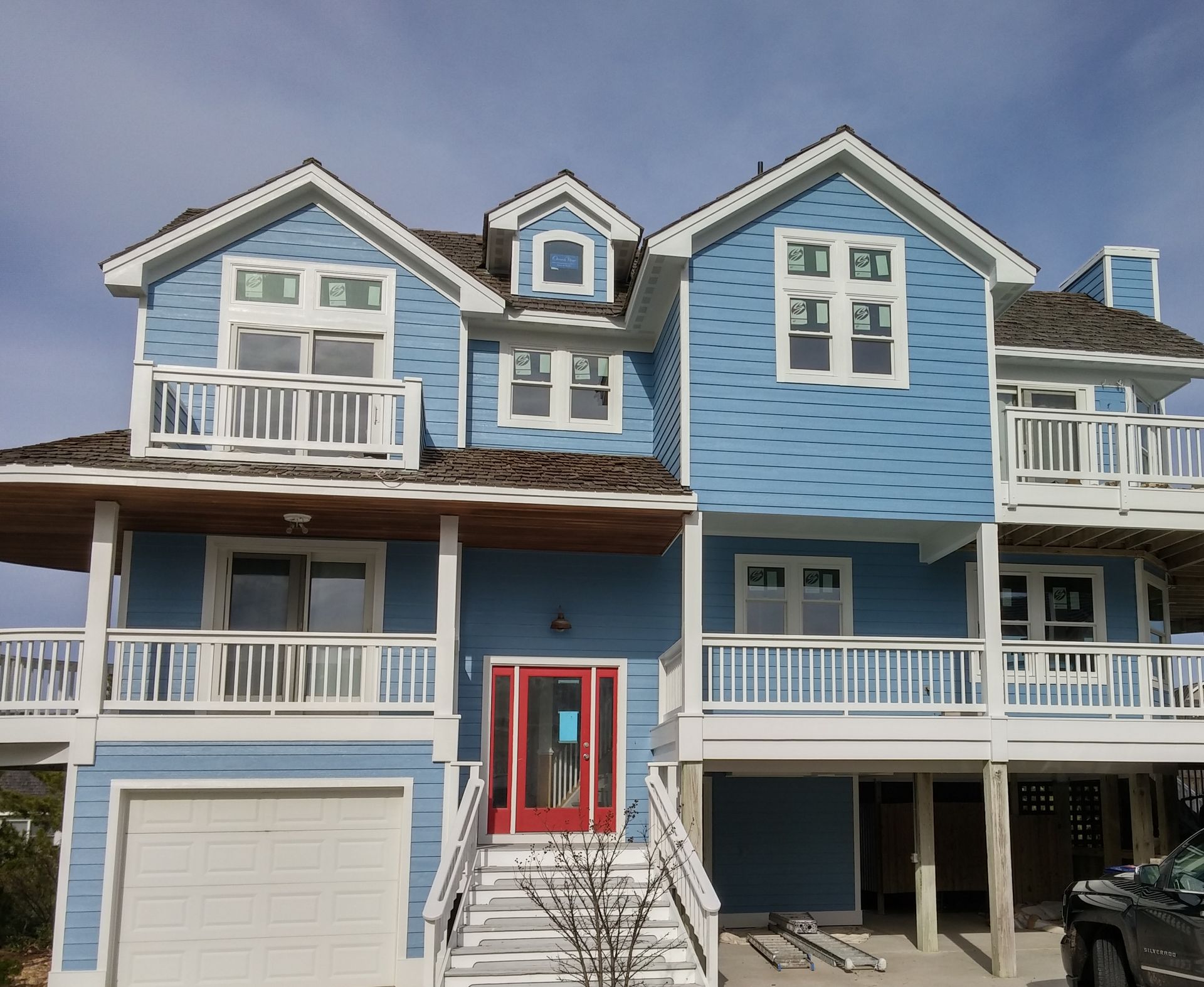 Blue two-story beach house with white trim, red door, and multiple balconies against a blue sky.