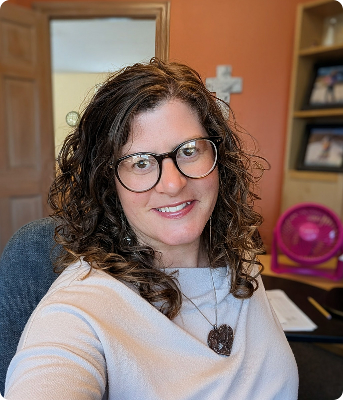 Woman with curly hair and glasses smiles. Interior, cross on wall, shelves with pictures visible.