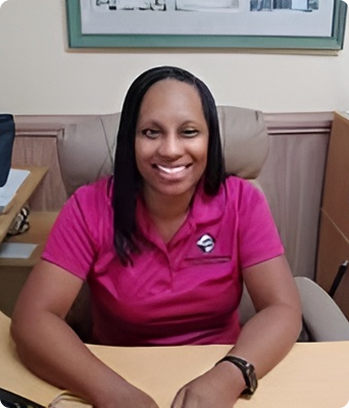 Woman with dark hair smiles, wearing a pink polo shirt, seated at a desk, with an office setting.