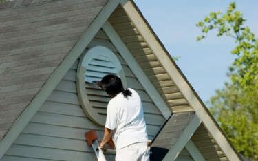 Person on a ladder painting a house's exterior siding near an oval vent.
