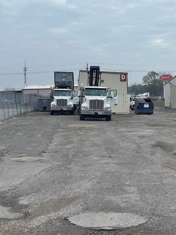 Two white semi trucks with containers on gravel lot in front of industrial buildings.