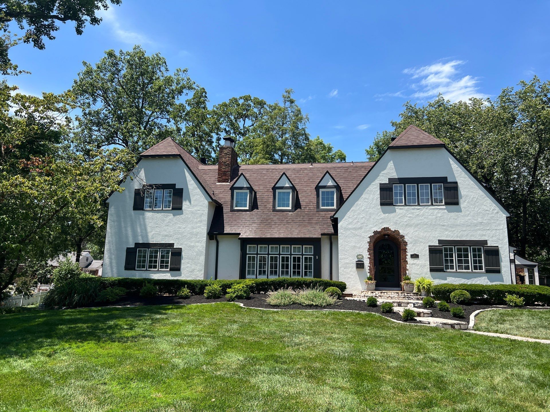 A two-story white house with dark trim and a brown shingled roof, set behind a lush green lawn under a bright blue sky.