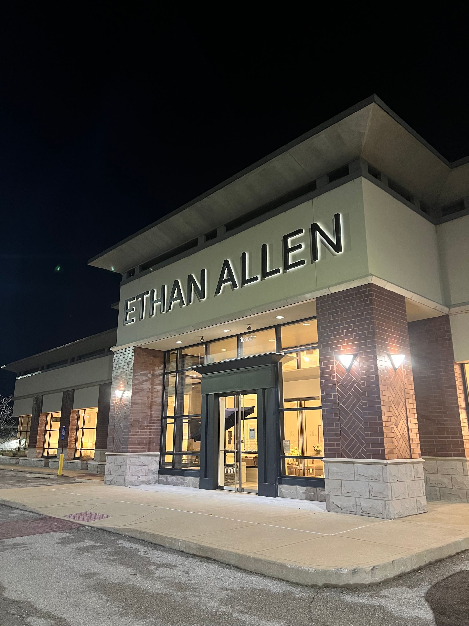 An Ethan Allen store front exterior at night, featuring a lit sign, brick accents, and large glass windows.