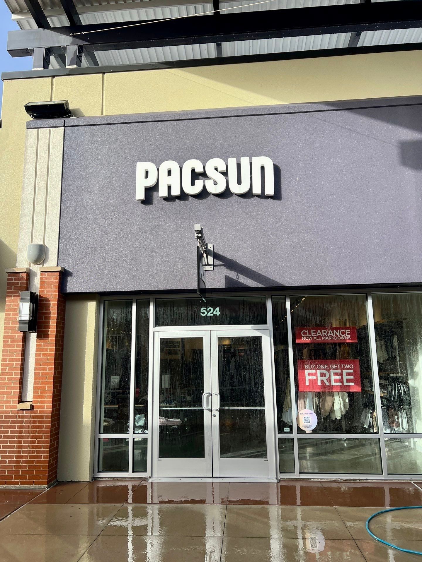 A PacSun retail store storefront with a gray facade, large glass doors, and window signs advertising a sale.