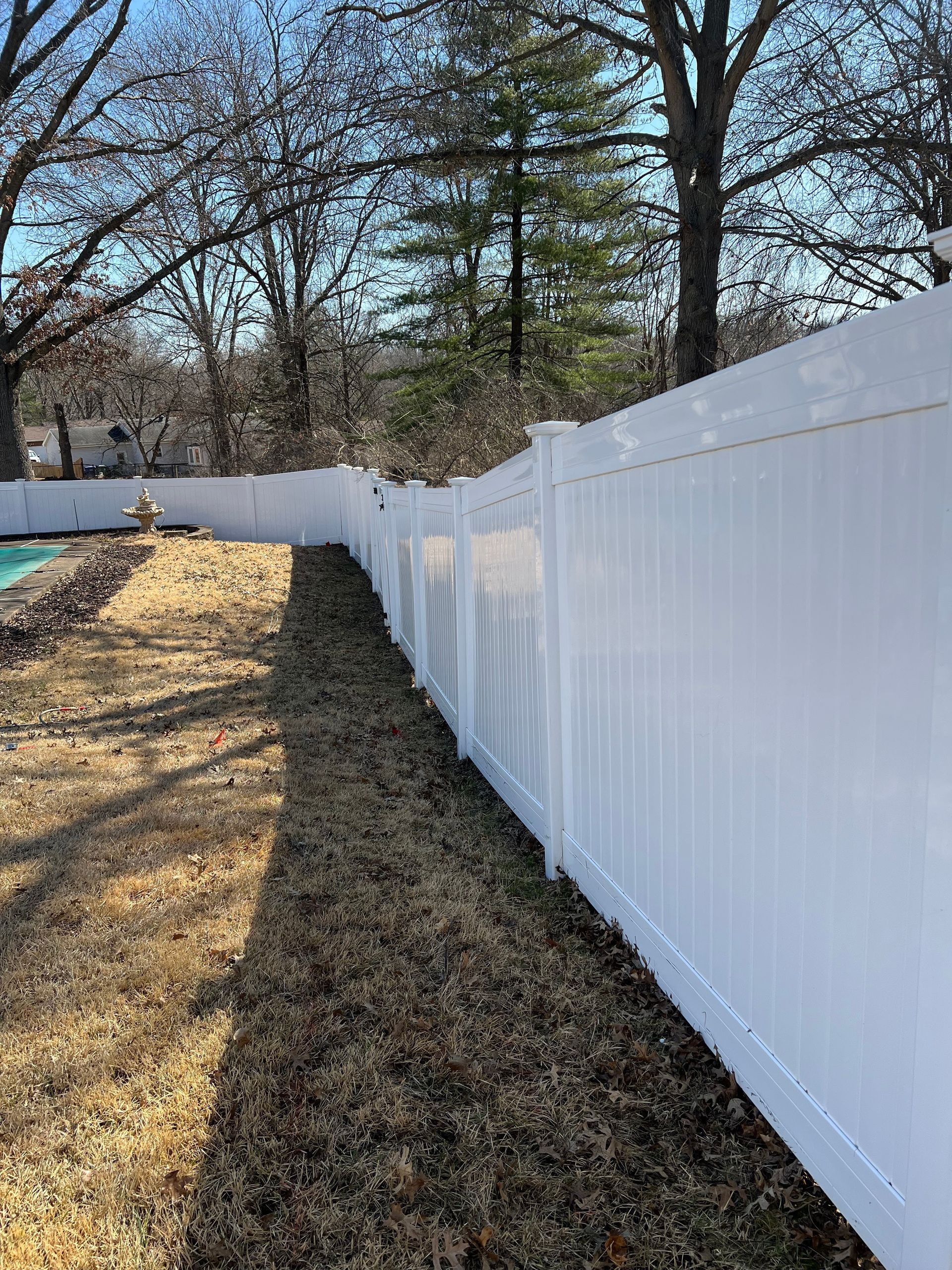 A white privacy fence runs along a grassy yard bordering a pool area under trees on a sunny day.
